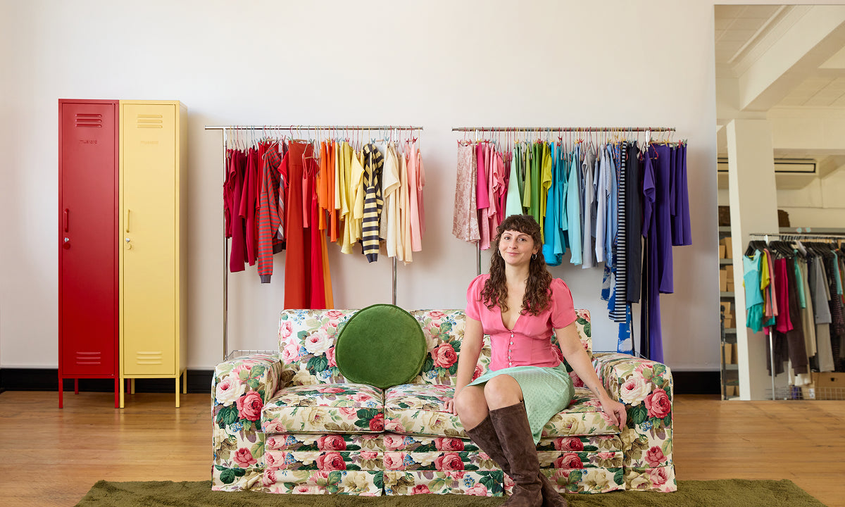 The Skinny lockers in Poppy red and Butter yellow beside rainbow-sorted garment racks in the Olga Joan fashion studio, founder seated on a floral sofa