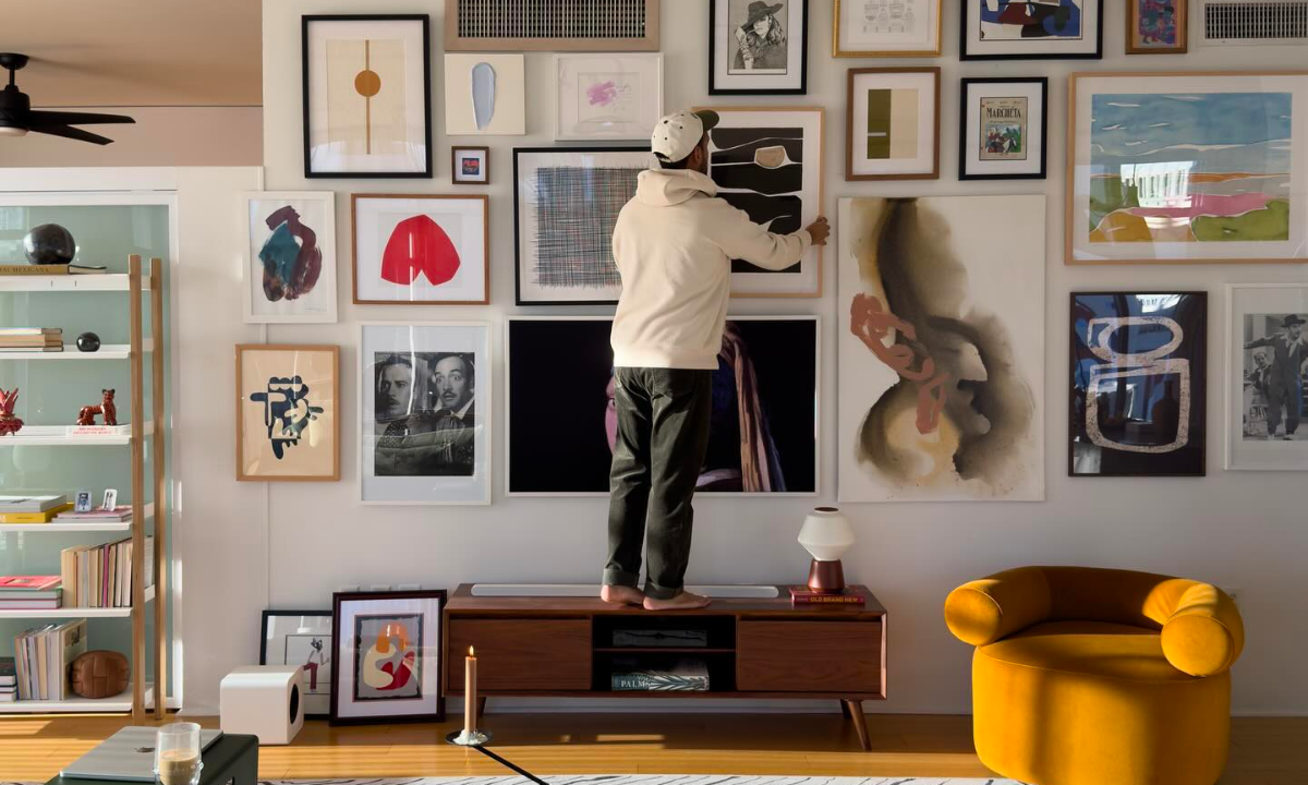 A man wearing a cream hoodie and black jeans is standing on top of a wooden console. He is adjusting an artwork situated in the centre of a gallery wall in his apartment.