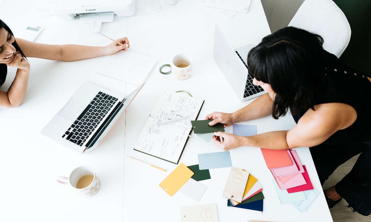 Becca leans over a white table to show Jess colour swatches