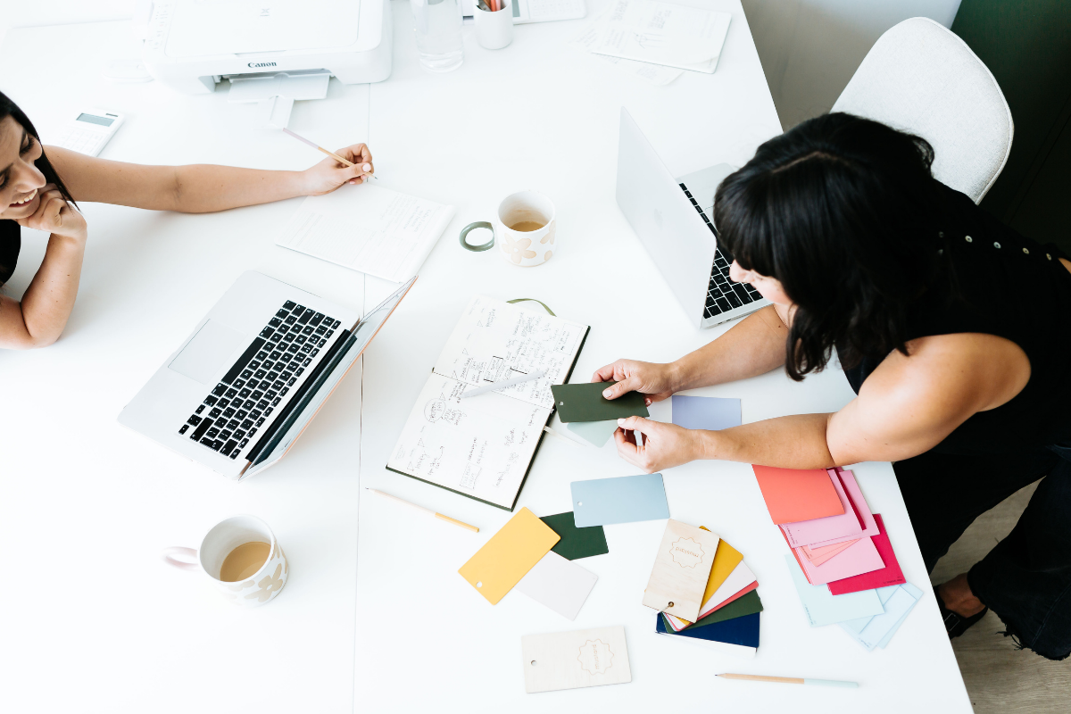 Becca leans over a white table to show Jess colour swatches
