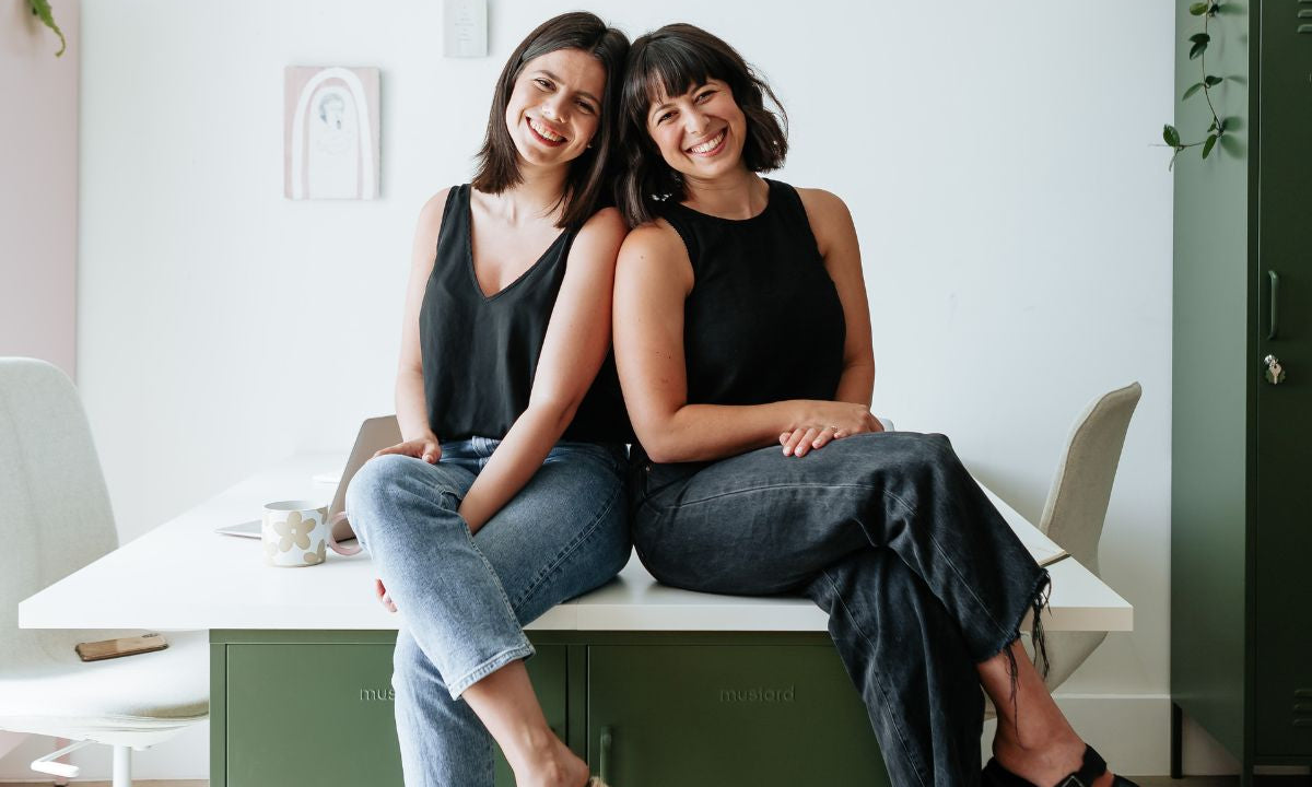 Mustard Made co-founders Becca and Jess sitting on a desk 