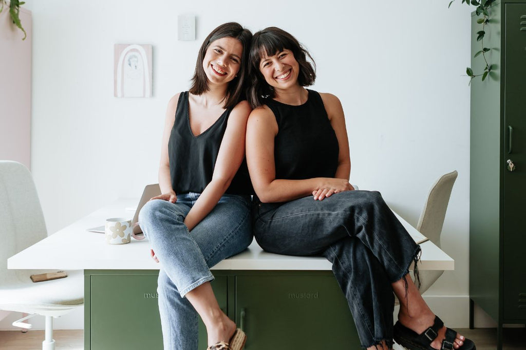 Mustard Made co-founders Becca and Jess sitting on a desk 