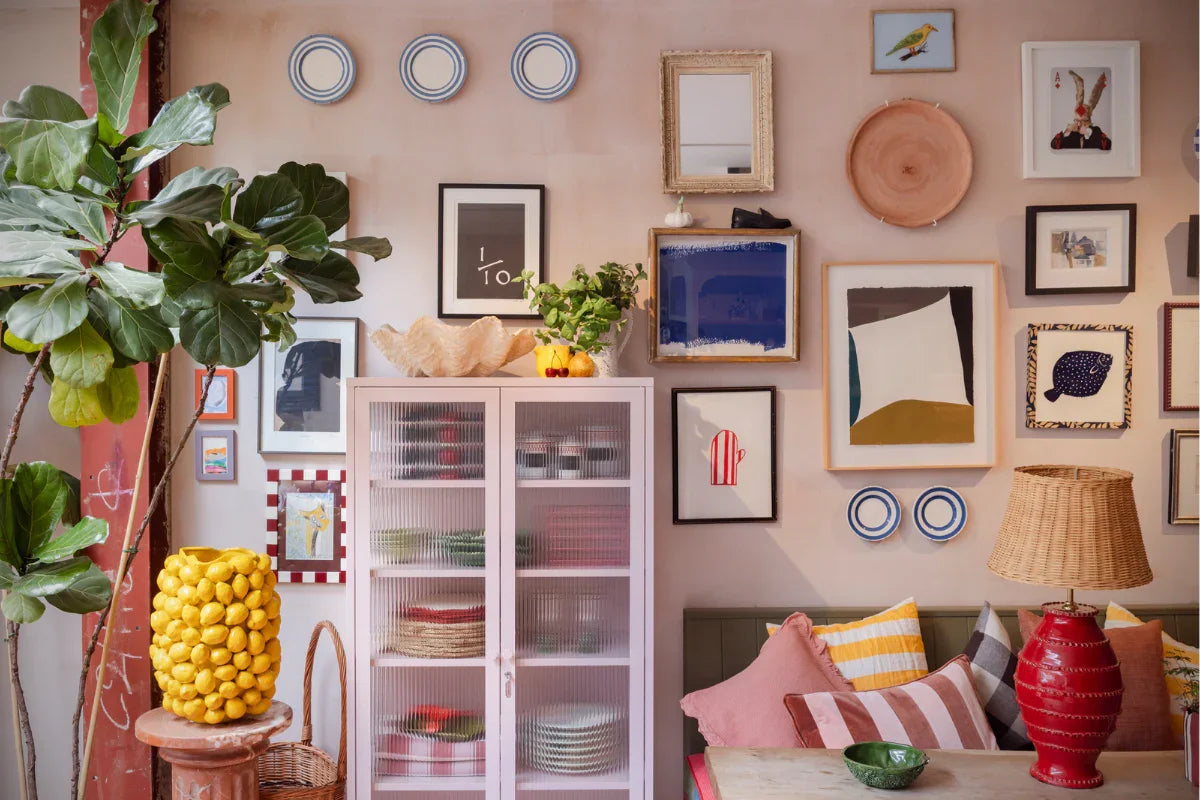 A Blush pink locker with glass doors against a pink wall filled with an eclectic mix of framed artworks, plants, cushions and other decor items.
