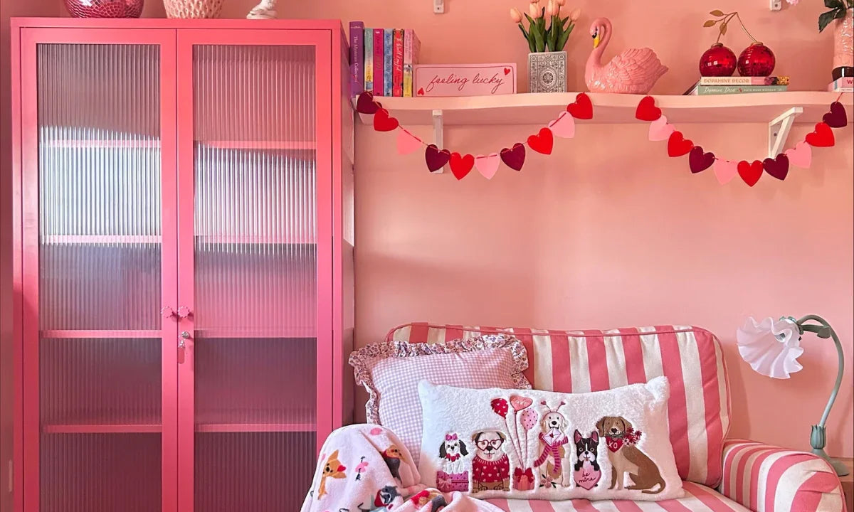 A pink-themed living room featuring a pink Mustard Made locker with fluted glass doors, a striped pink and white arm chair and two floating shelves displaying knick-knacks.