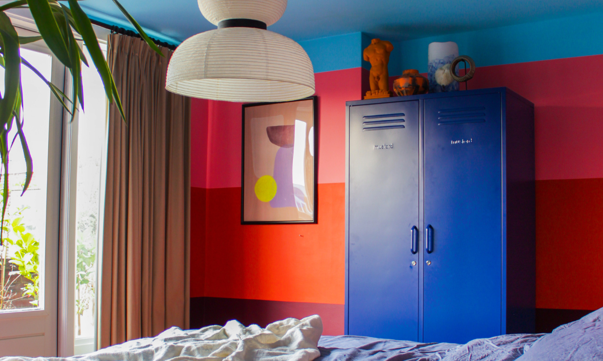 A bedroom is painted in thick horizontal stripes of plum, red and pink, with a light blue ceiling. There is a Navy Twinny locker next to an abstract print.