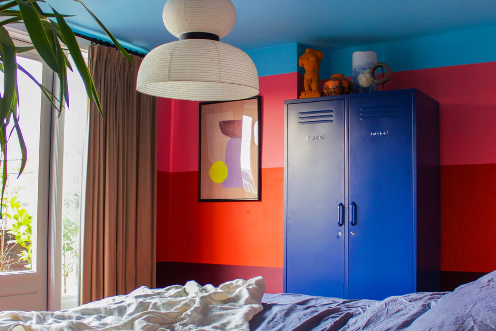 A bedroom is painted in thick horizontal stripes of plum, red and pink, with a light blue ceiling. There is a Navy Twinny locker next to an abstract print.