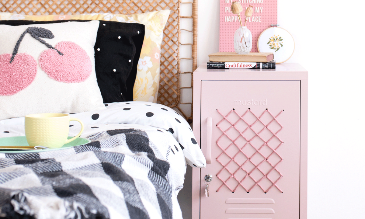 A short blush locker featuring a cross-stitch patterned door is styled next to a rattan bed with a mix of patterned linens and cushions.