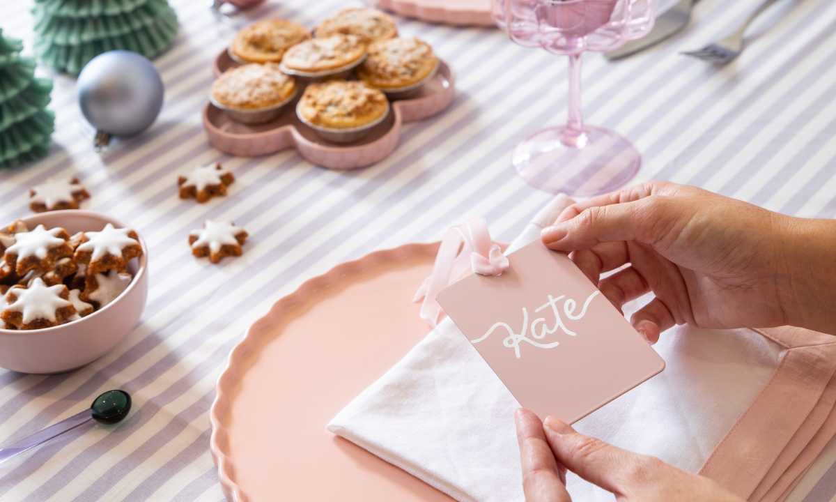 A pastel coloured tablescape features star-shaped gingerbread, Christmas tree candles and pink glassware. Someone is holding a Blush Mustard Made swatch which has been crafted into a place card with the name Kate written on it.