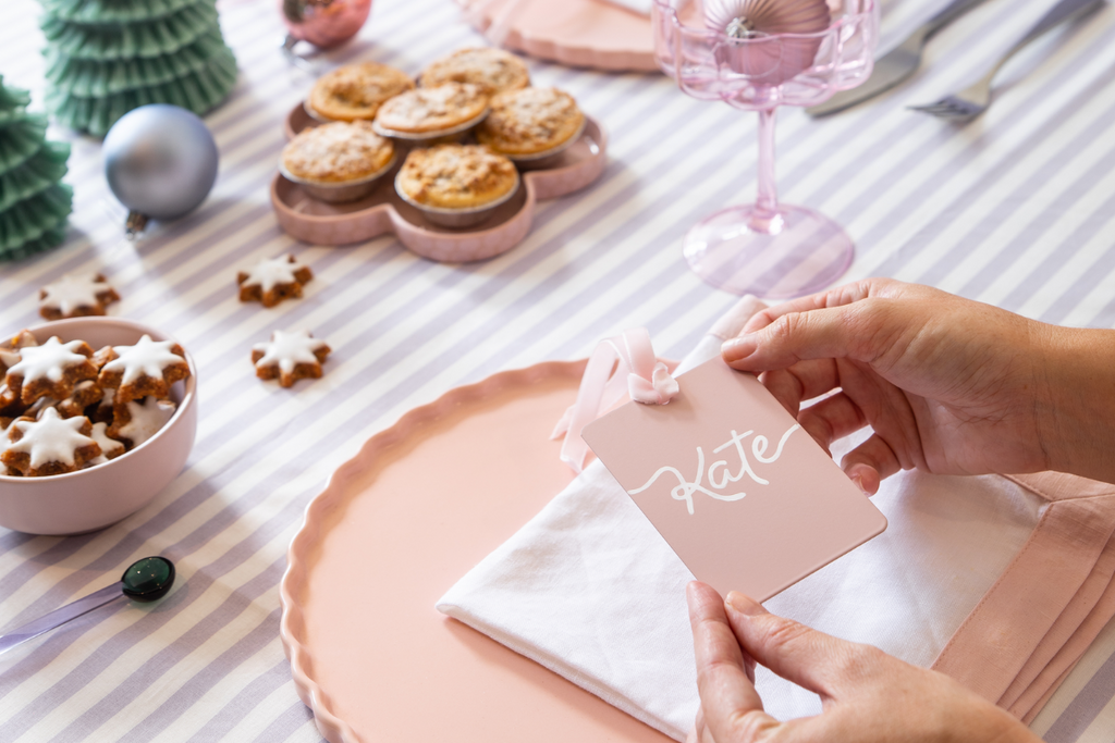 A pastel coloured tablescape features star-shaped gingerbread, Christmas tree candles and pink glassware. Someone is holding a Blush Mustard Made swatch which has been crafted into a place card with the name Kate written on it.