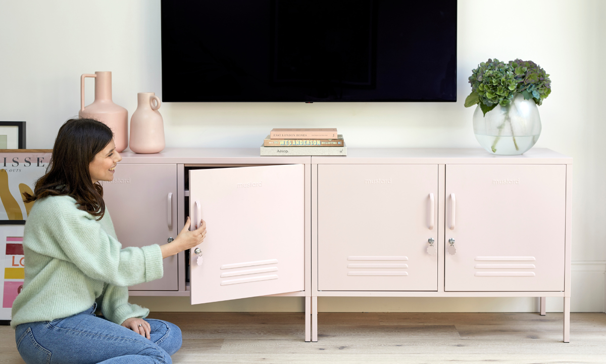 Jess kneels in front of two Blush Lowdown lockers. She is wearing blue jeans and a pale green top, and there is a TV mounted on the wall behind her.