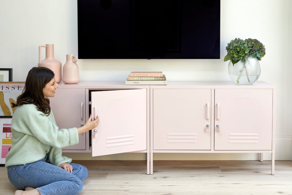 Jess kneels in front of two Blush Lowdown lockers. She is wearing blue jeans and a pale green top, and there is a TV mounted on the wall behind her.