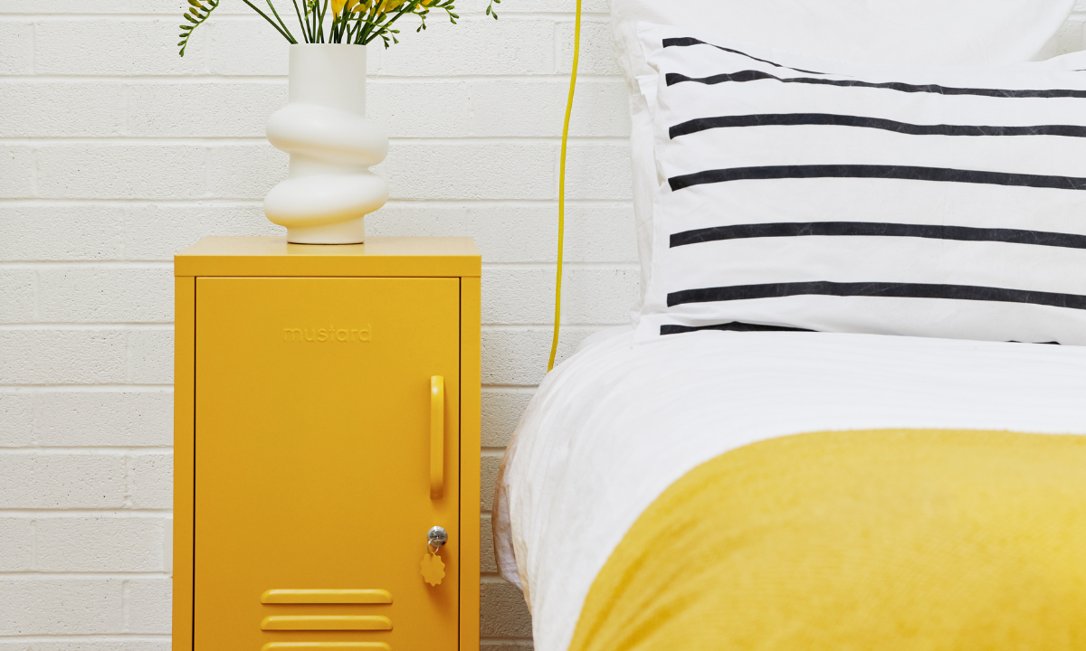 A Mustard yellow Shorty locker sits next to a bed dressed in Mustard linens with a black and white striped pillow. There is a vase of yellow flowers on top of the locker.