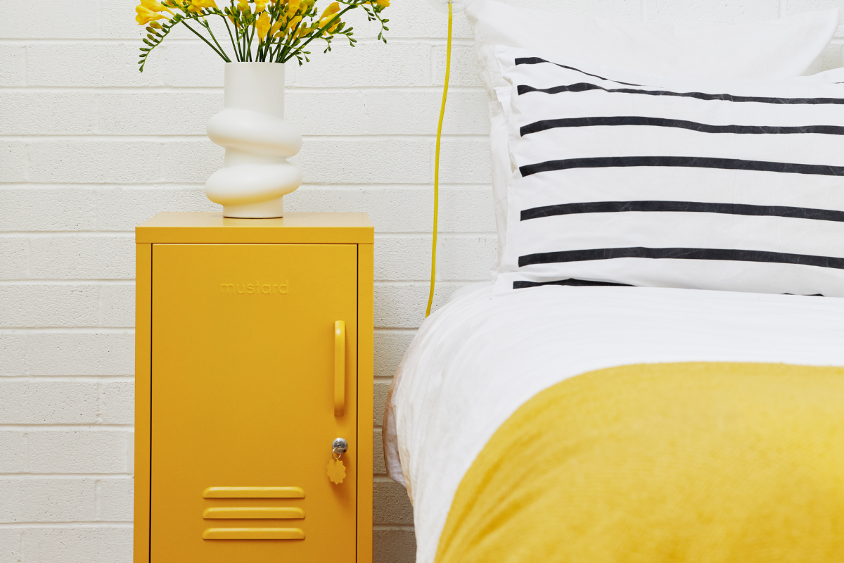 A Mustard yellow Shorty locker sits next to a bed dressed in Mustard linens with a black and white striped pillow. There is a vase of yellow flowers on top of the locker.