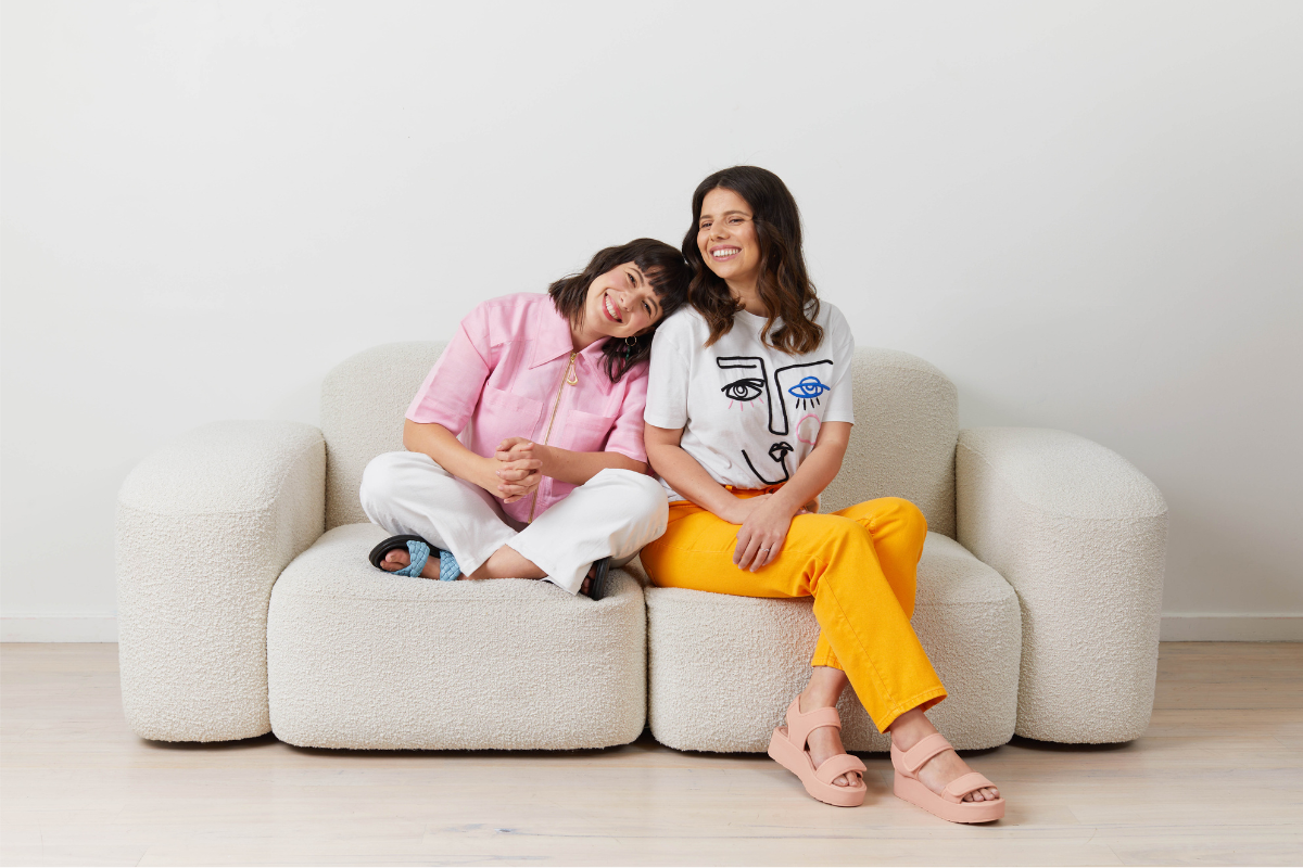 Two white women with dark hair sit on a cream boucle couch. Becca is wearing a pink shirt and smiling with her head on Jess' shoulder.