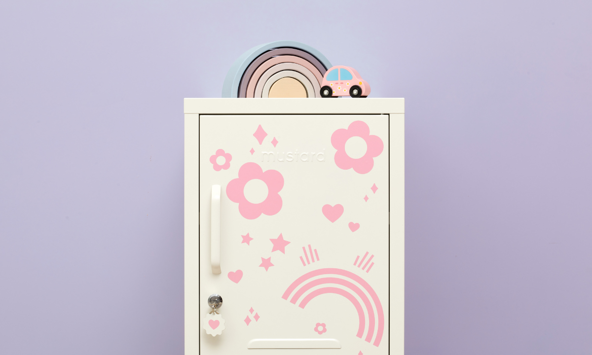 A small white locker is covered in pink decals in various shapes including flowers, rainbows and hearts. There is a pastel wooden rainbow and a wooden toy car on top.