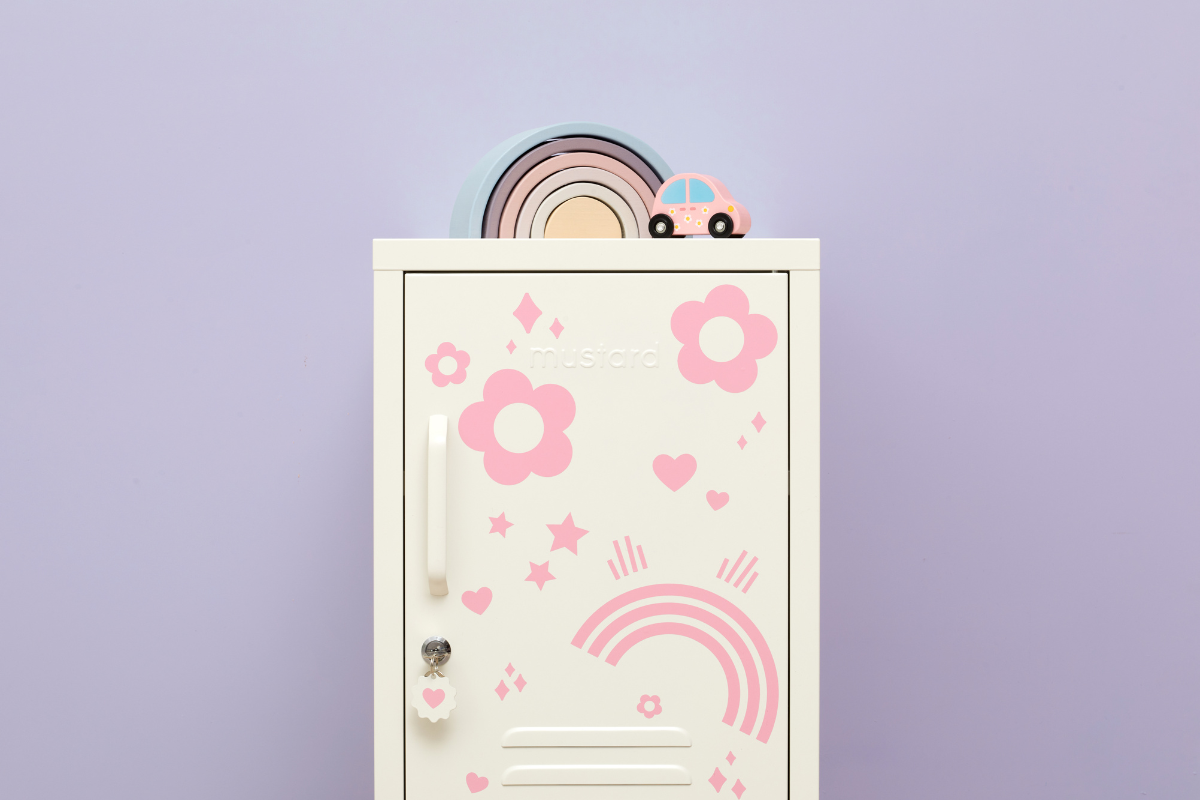 A small white locker is covered in pink decals in various shapes including flowers, rainbows and hearts. There is a pastel wooden rainbow and a wooden toy car on top.