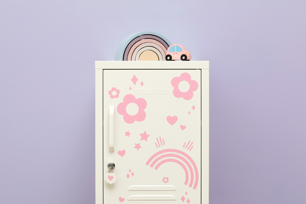 A small white locker is covered in pink decals in various shapes including flowers, rainbows and hearts. There is a pastel wooden rainbow and a wooden toy car on top.