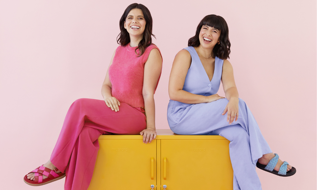 Becca and Jess sit on The Lowdown in Mustard. Jess is wearing a bright, berry-coloured pink knitted singlet and long pants. Becca wears a light, lilac purple vest and long pants.