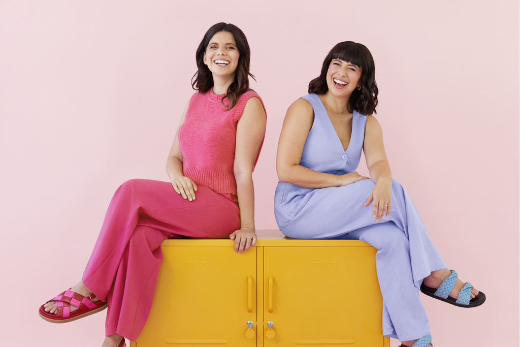 Becca and Jess sit on The Lowdown in Mustard. Jess is wearing a bright, berry-coloured pink knitted singlet and long pants. Becca wears a light, lilac purple vest and long pants.