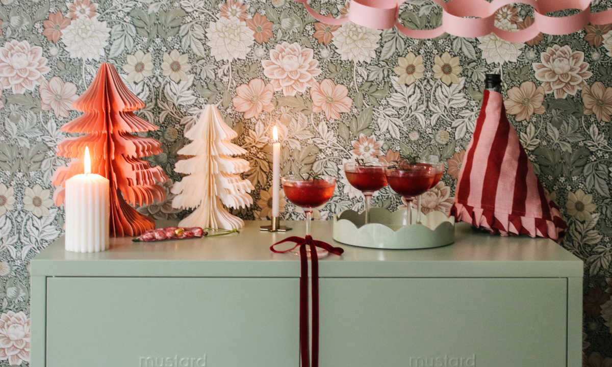 The top of a Sage Mustard Made locker is decorated with red and pink Christmas decorations and lit candles. There is pink and green floral wallpaper behind it with a pink paper chain streamer.