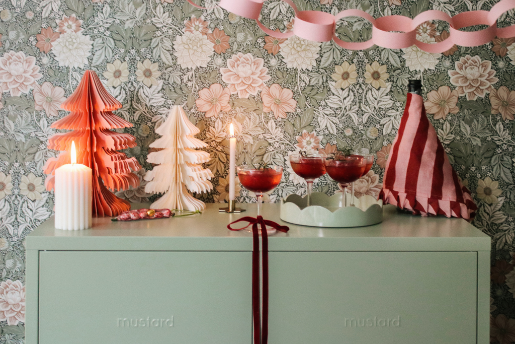 The top of a Sage Mustard Made locker is decorated with red and pink Christmas decorations and lit candles. There is pink and green floral wallpaper behind it with a pink paper chain streamer.