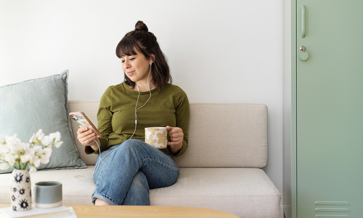 Becca sits on a cream couch next to a Sage Skinny locker. She is holding a mug and listening to a podcast with headphones.