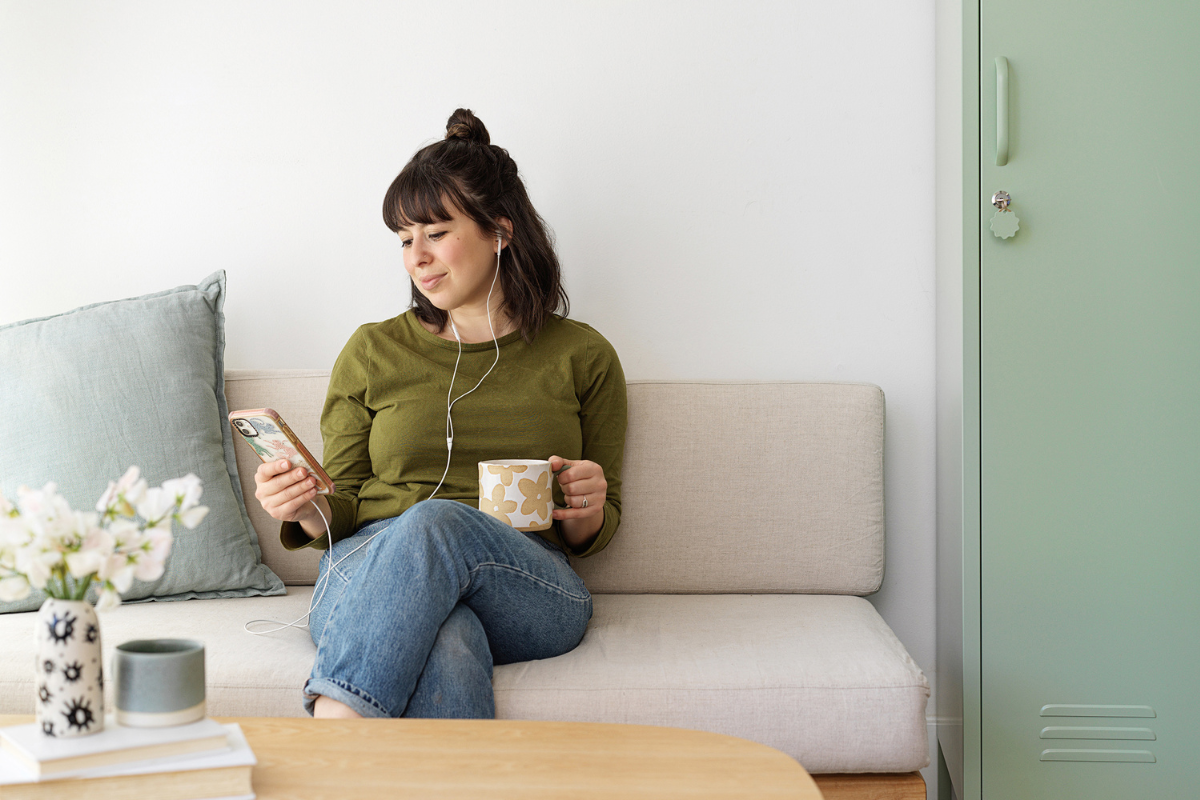Becca sits on a cream couch next to a Sage Skinny locker. She is holding a mug and listening to a podcast with headphones.
