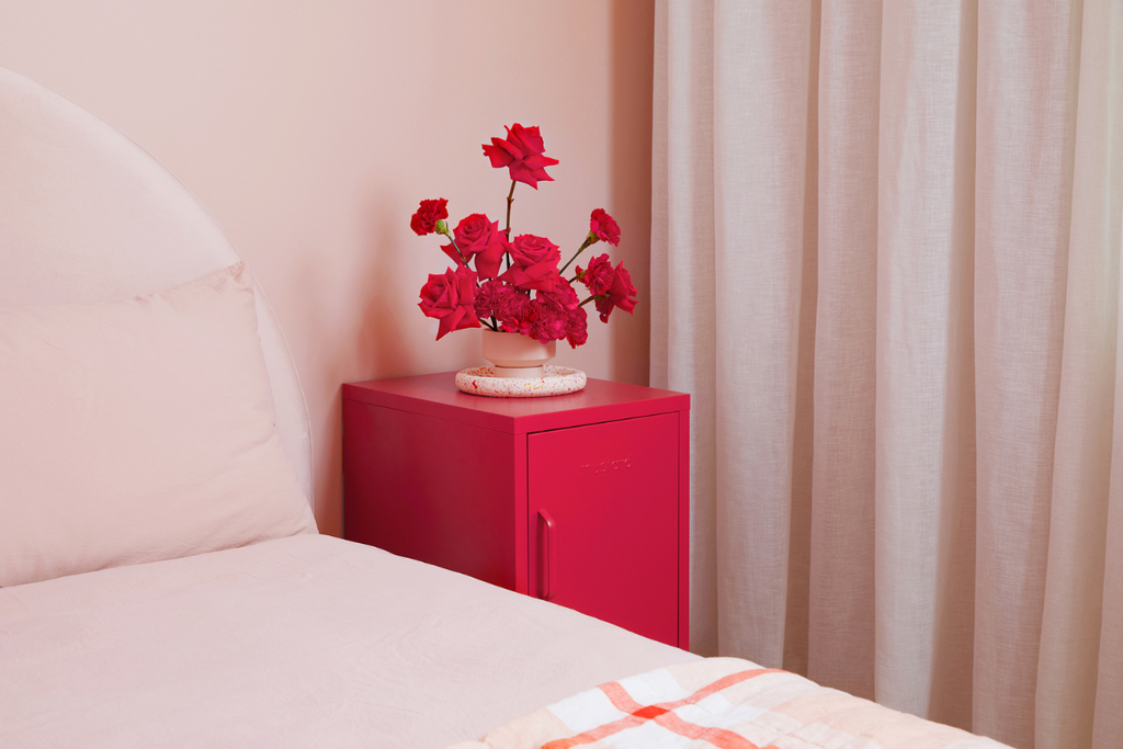 The Shorty in Poppy next to a bed, with a vase of red roses on top