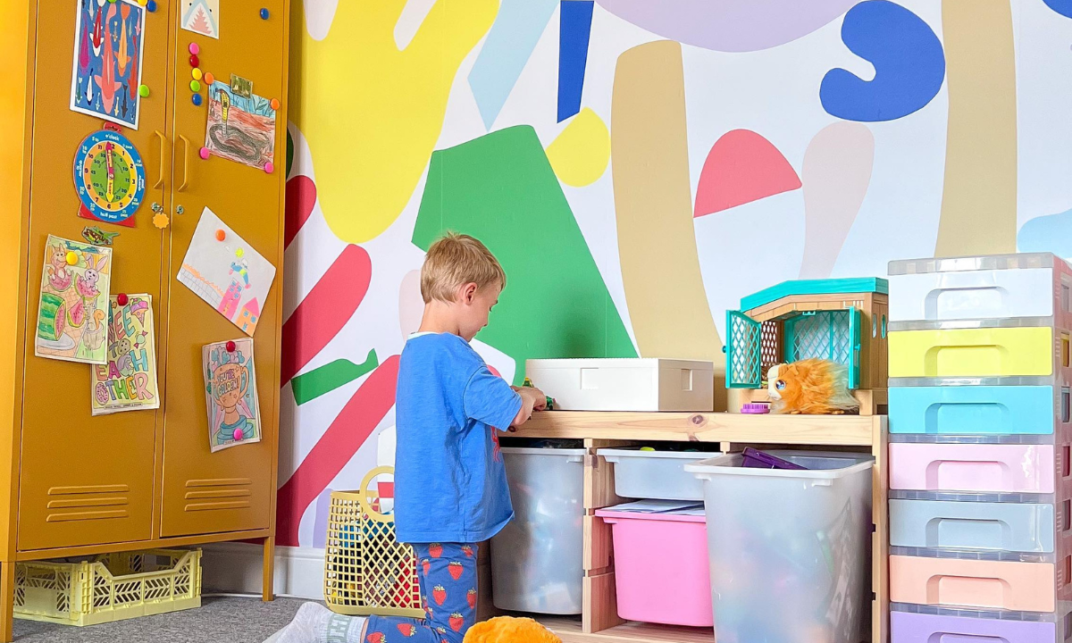 A child kneels in front of a colourful activity zone filled with tubs and trays of toys. There is a Mustard Twinny behind them covered in kids art and magnets, and the wall is decorated with bold, bright geometric shapes.