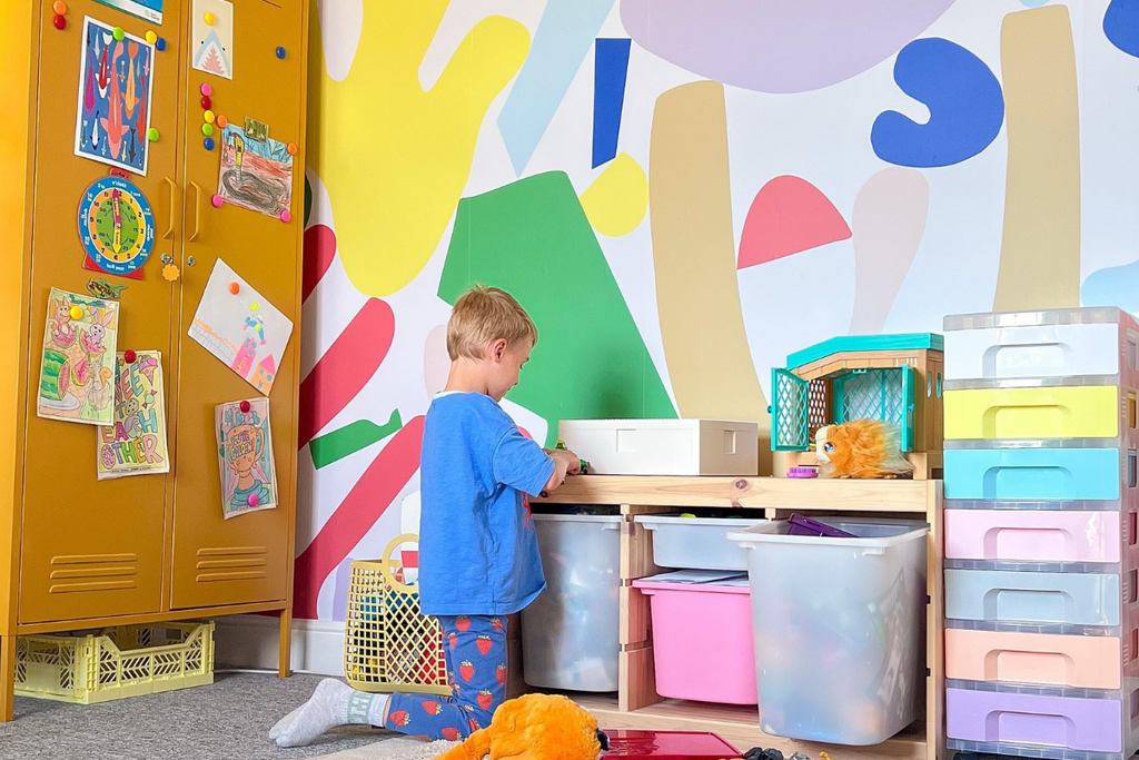 A child kneels in front of a colourful activity zone filled with tubs and trays of toys. There is a Mustard Twinny behind them covered in kids art and magnets, and the wall is decorated with bold, bright geometric shapes.