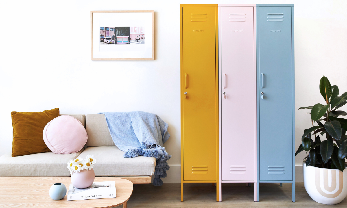 Three Mustard Made Skinny lockers stand side by side. From the left, the first is Mustard, then Blush then Ocean. To the left is a sofa with a wooden, timber frame, beige padding and cushions of various shapes and colours sit on top. 
