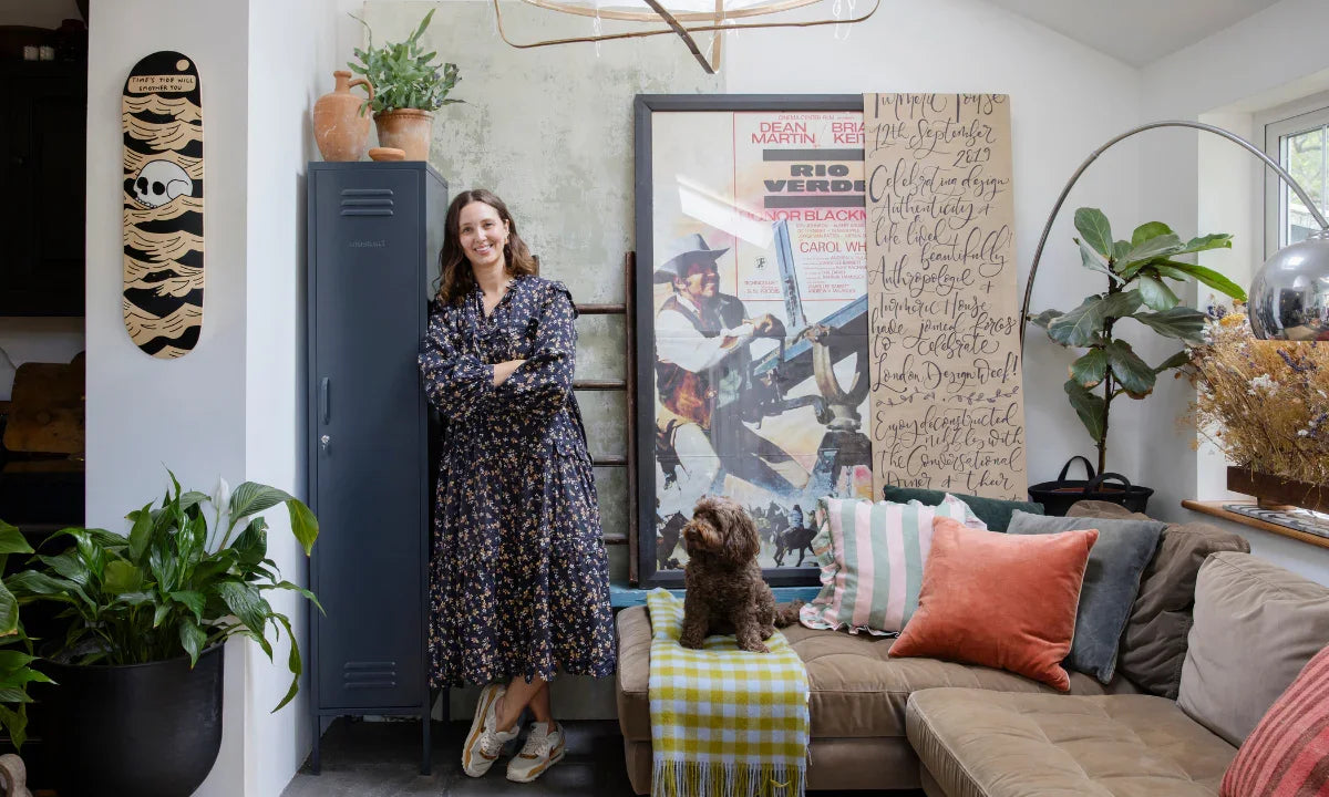 Clare Watson wears a flowing floral dress and sneakers as she leans against a Slate Skinny Mustard Made locker. She is surrounded by plants, artworks and throw cushions in her living room.