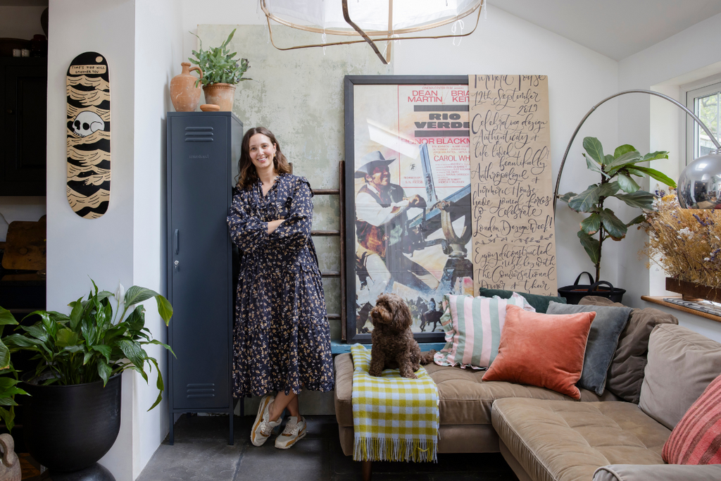 Clare Watson wears a flowing floral dress and sneakers as she leans against a Slate Skinny Mustard Made locker. She is surrounded by plants, artworks and throw cushions in her living room.